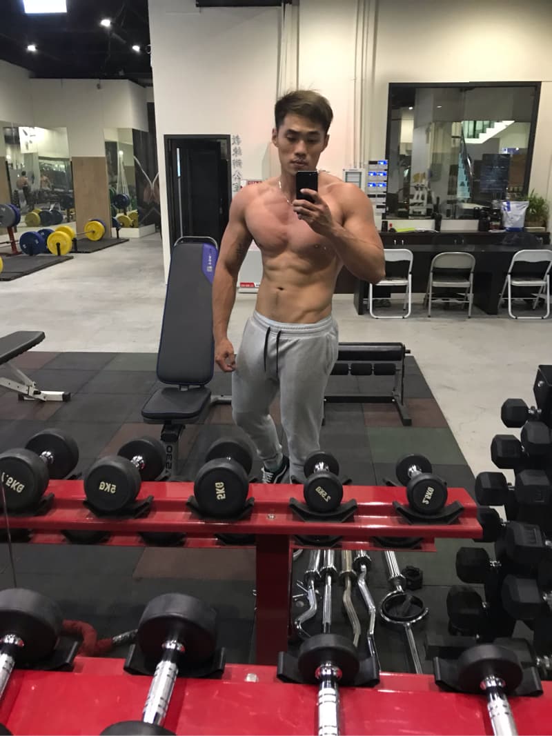 這張「健身房」的照片是一個男子在鏡子前自拍，他穿著灰色的運動褲，展現了他的腹肌。照片的背景是健身房，可以看到一些健身器材，像是啞鈴和器械。