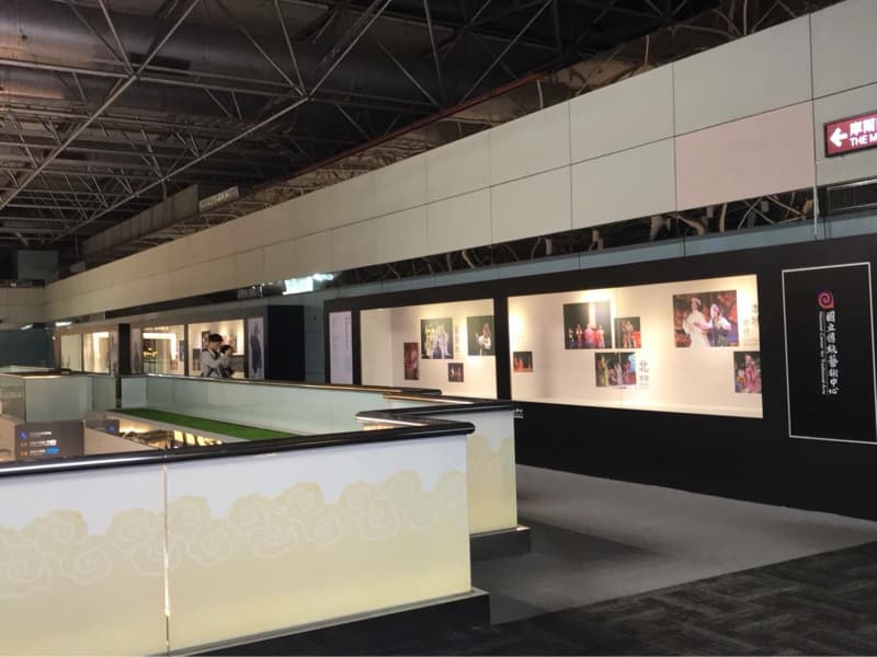 這張「藝術展覽」的照片是一個展覽空間，牆上展示著許多中國戲劇的劇照，還有展覽的介紹文字。空間中有步道、欄杆、草皮以及指示牌。