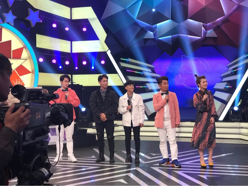 這張照片是五個藝人在舞台上錄製節目的照片。 他們都穿著時尚的服裝，並且正在說話或唱歌。  後方是舞台背景，看起來像是節目製作的佈景。