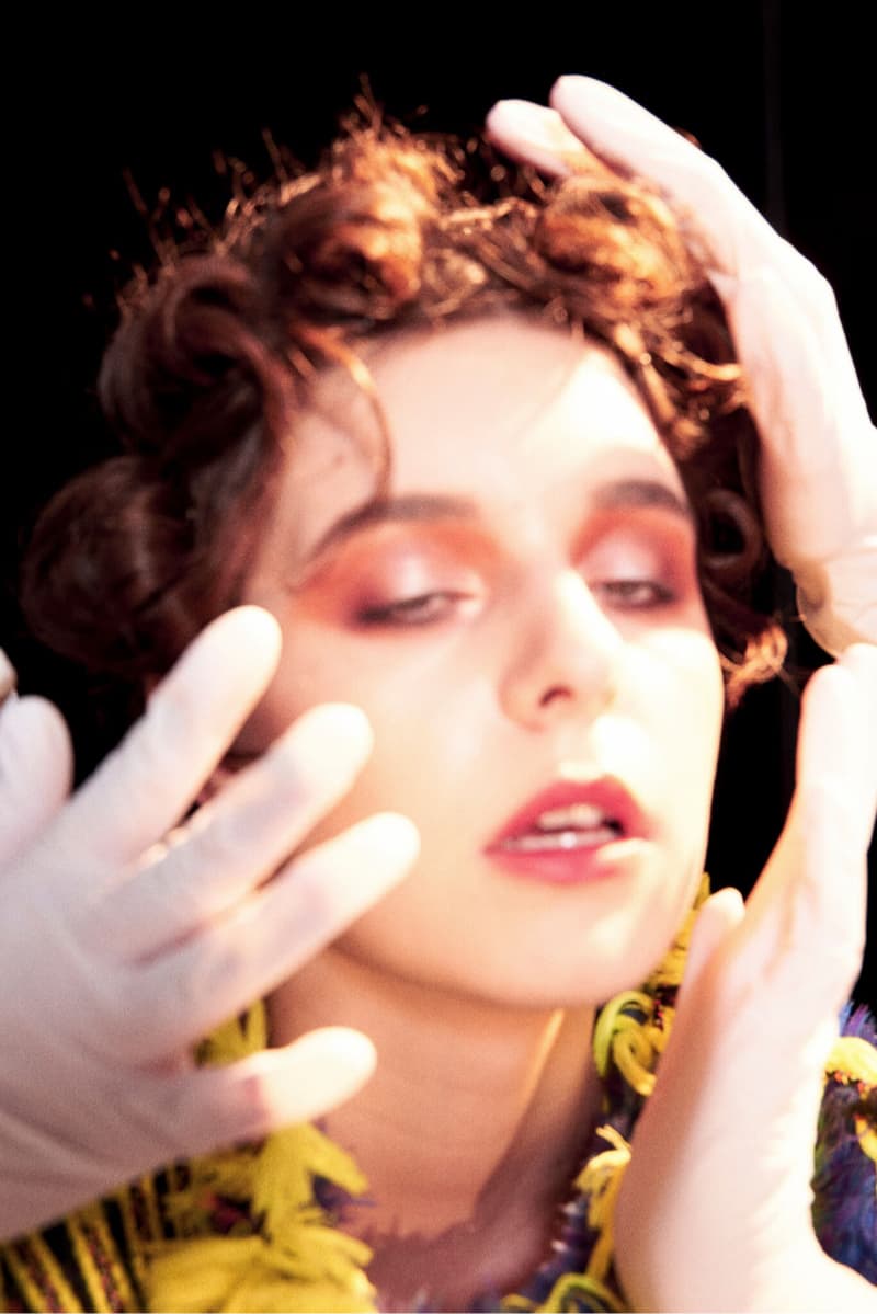 這張照片是一個女性，她戴著金色和藍色的飾品，手上戴著白色的手套，她的臉被手套遮住，並用粉紅色和橘色眼影化妝。