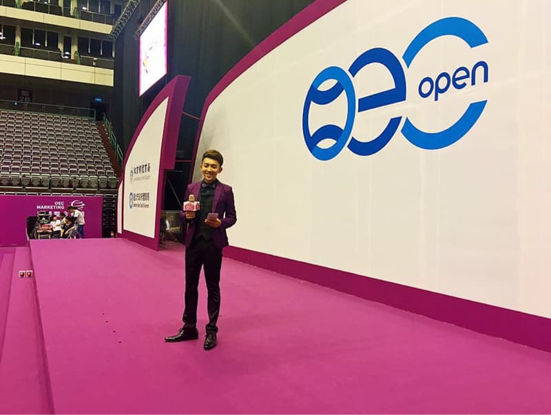 這張「網球比賽」的照片是，一位主持人站在粉紅色的舞臺上，背景是比賽的標誌和觀眾席。