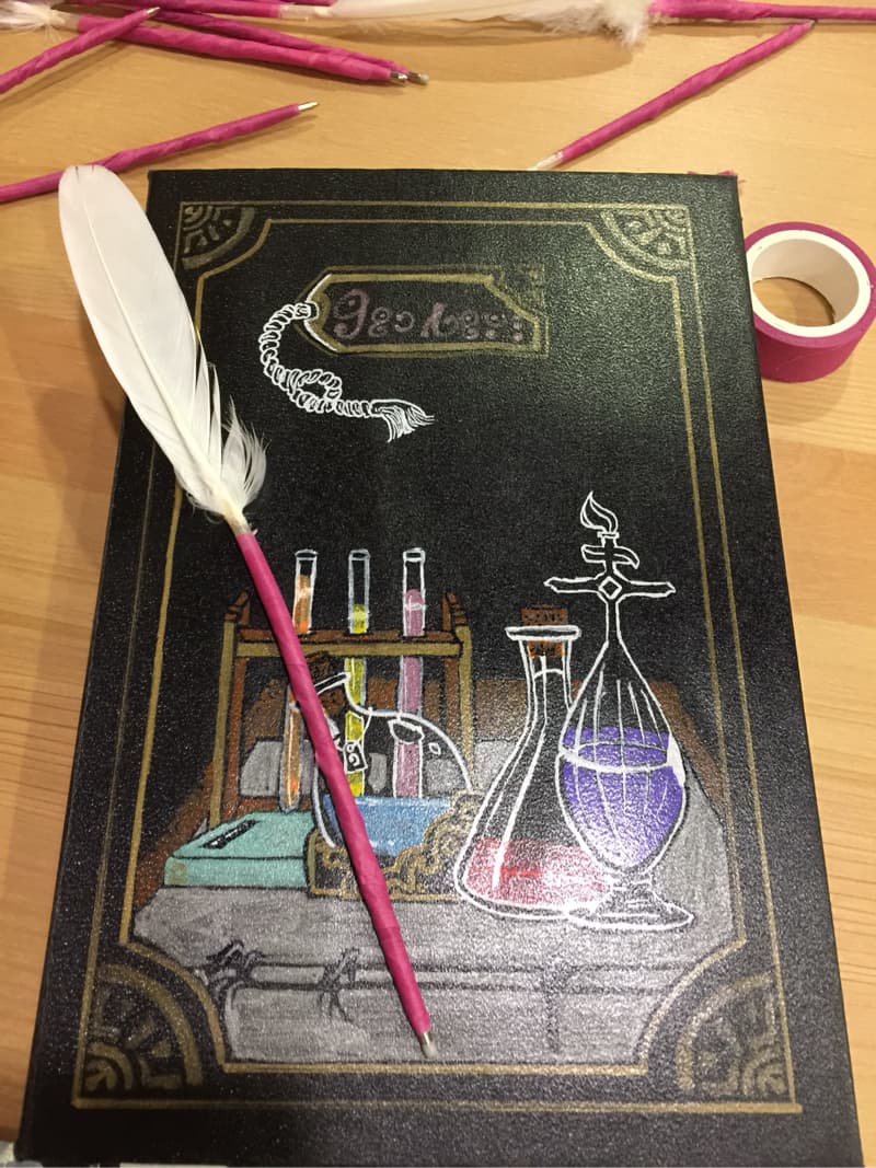 這張照片是一個黑底金邊的畫布，上面畫著一個瓶子，裡面裝著紫色的液體。瓶子旁邊還有一個試管架，裡面放著試管。畫布上還有一根羽毛筆。