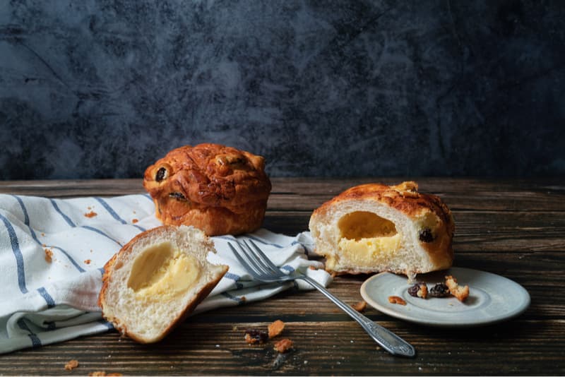 這張「麵包」的照片是兩顆奶油麵包，其中一顆被切開，露出了裡面的奶油餡。旁邊還有一塊切下來的麵包、一個餐盤、一根叉子和一塊條紋布。