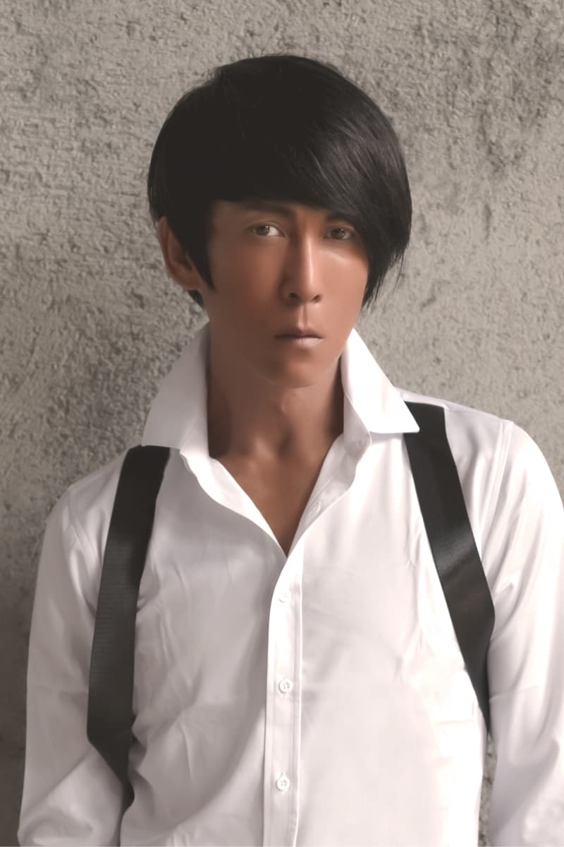 這張照片是一個穿著白色襯衫的年輕男子。他表情嚴肅，頭髮烏黑，眼神堅定，他的肩膀上有一條黑色的吊帶，整體造型非常酷。