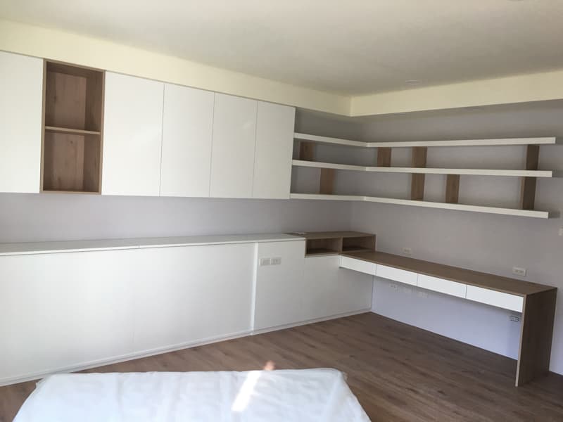 這張照片是一個現代化的臥室，房間有一個木質的書桌，書桌的下方有兩個抽屜，書桌旁邊有一個白色床頭櫃，牆上有一個白色的層板架，房間的整體風格非常簡約。