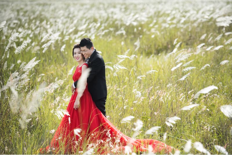 這張照片是一對情侶在一片草地上擁抱。他們穿著紅色禮服和黑色西裝，看起來非常甜蜜。他們周圍的草地上長滿了白色的芒草，營造出一種浪漫的氛圍。