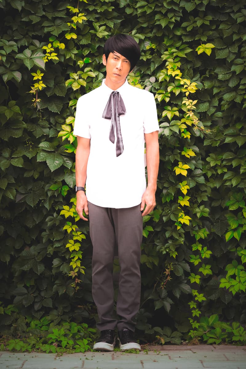 這張照片是一個穿著白襯衫和黑褲子的年輕男子站在綠色藤蔓牆面前。