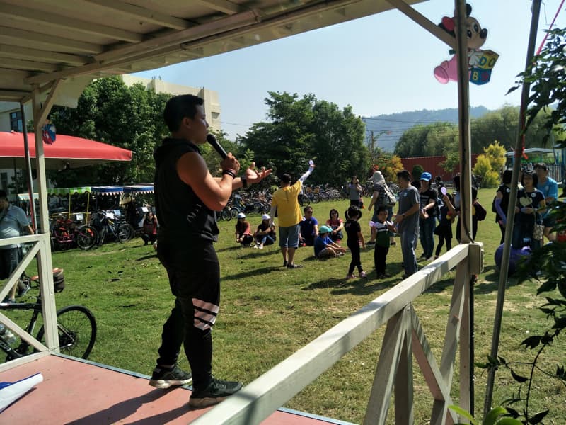 這張「戶外活動」的照片是一群人聚在一起，其中一位站在臺上用麥克風講話，看起來像是在主持活動。舞臺前有一群人聆聽，旁邊還有自行車和綠色的草地。