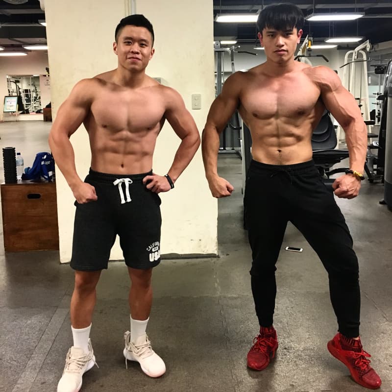 這張照片是兩個年輕男子在健身房的鏡子前擺姿勢，他們都展示著健美的肌肉和身材。