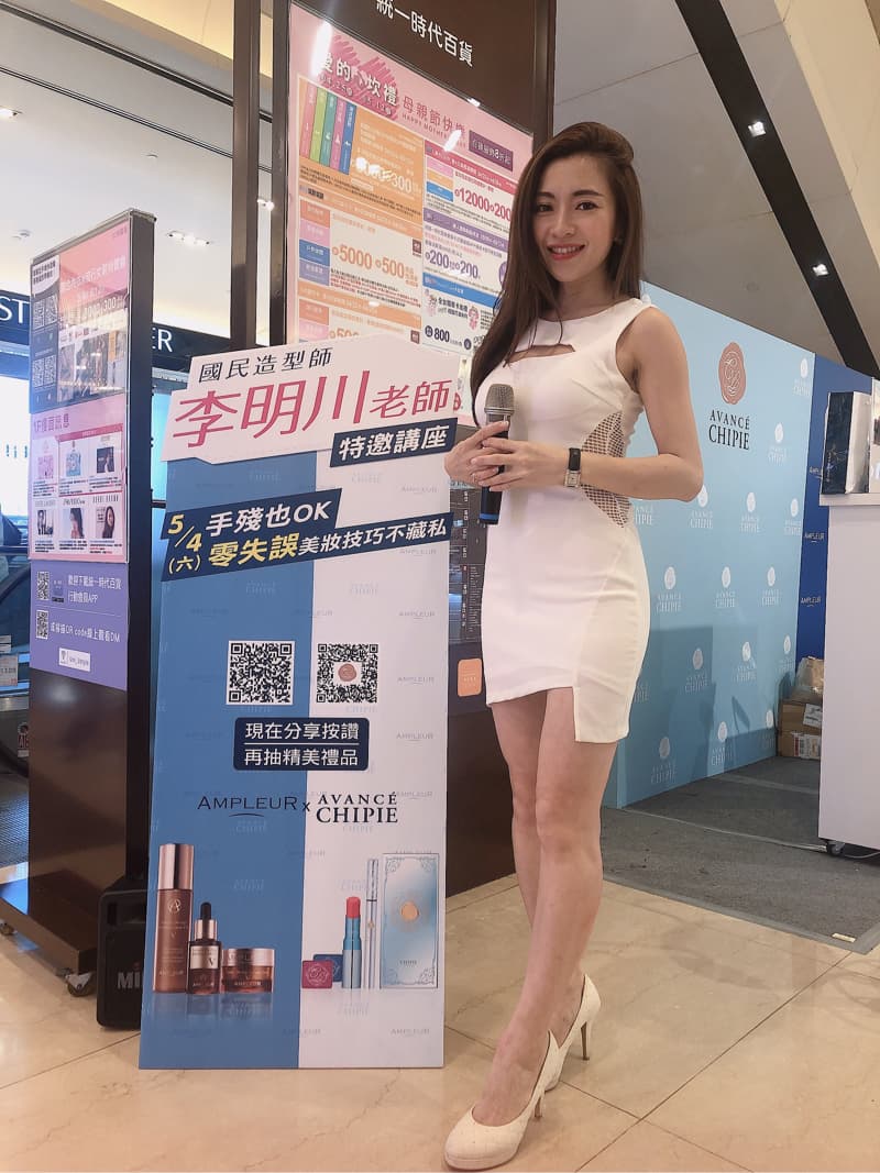 這張「美容產品宣傳」的照片是一個穿著白色連衣裙的女性，她站在一個廣告看板前，看板上展示了美容產品和品牌資訊，女性手持麥克風，似乎正在進行產品介紹。