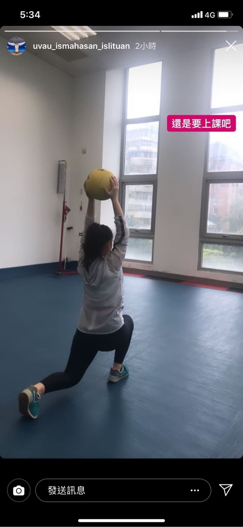 這張照片是一個女人在室內健身房裡做運動，她正在做一個舉重運動，右手高高舉起一個黃色的藥球，左腳伸直，右腳彎曲，身體微微前傾，看起來很努力。