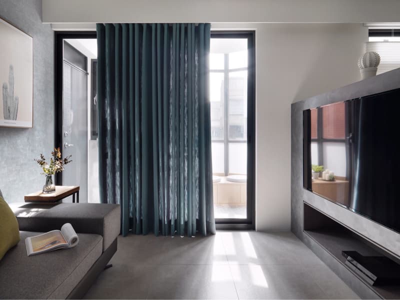 這張客廳的照片是一張灰色沙發，旁邊放著一張小茶几，茶几上擺放著一束花。沙發旁邊是深藍色的窗簾，窗戶外可以看到陽臺。牆上裝了一個電視機，電視機下方有一個黑色電視櫃。