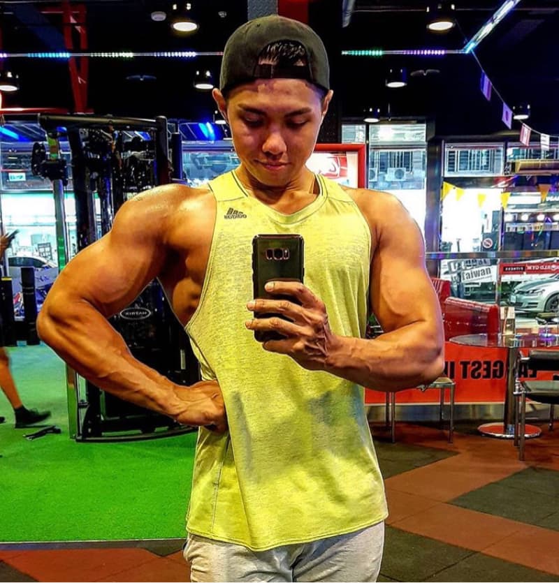 這張「健身房自拍」的照片是一個年輕男子在健身房裡自拍。他穿著黃色的背心和灰色的短褲，戴著黑色的帽子。他正在用手機自拍，展示他強壯的肌肉。他站在健身房的鏡子前，背景是健身器材和一些桌子。