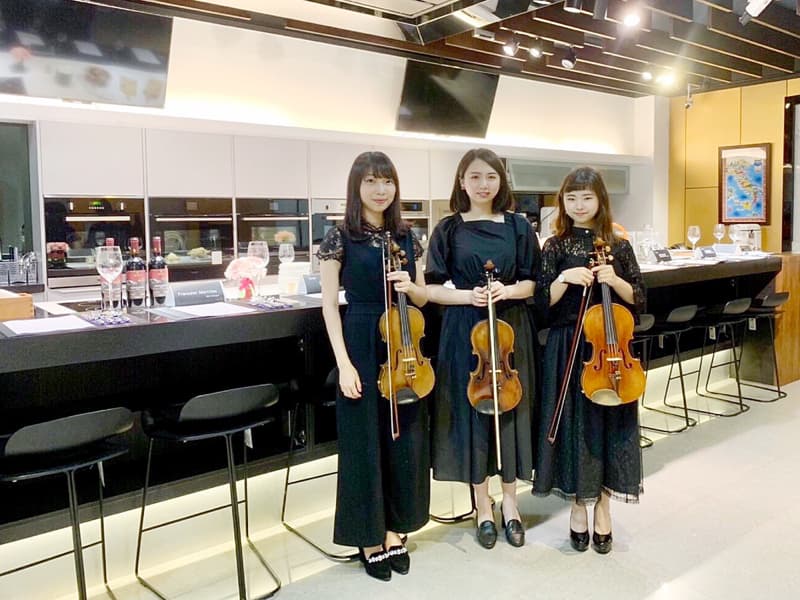 這張照片是三個穿著黑色連衣裙的女性站在廚房吧檯前，她們每個人都拿著小提琴。
