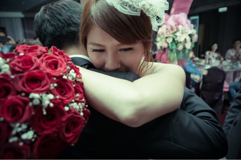 這張照片是一對新人擁抱的畫面，新娘將頭埋在新郎的肩上，看起來幸福而甜蜜。他們身穿婚禮服裝，新娘的頭上戴著頭紗，周圍還有紅色玫瑰花，營造出溫馨浪漫的氣氛。