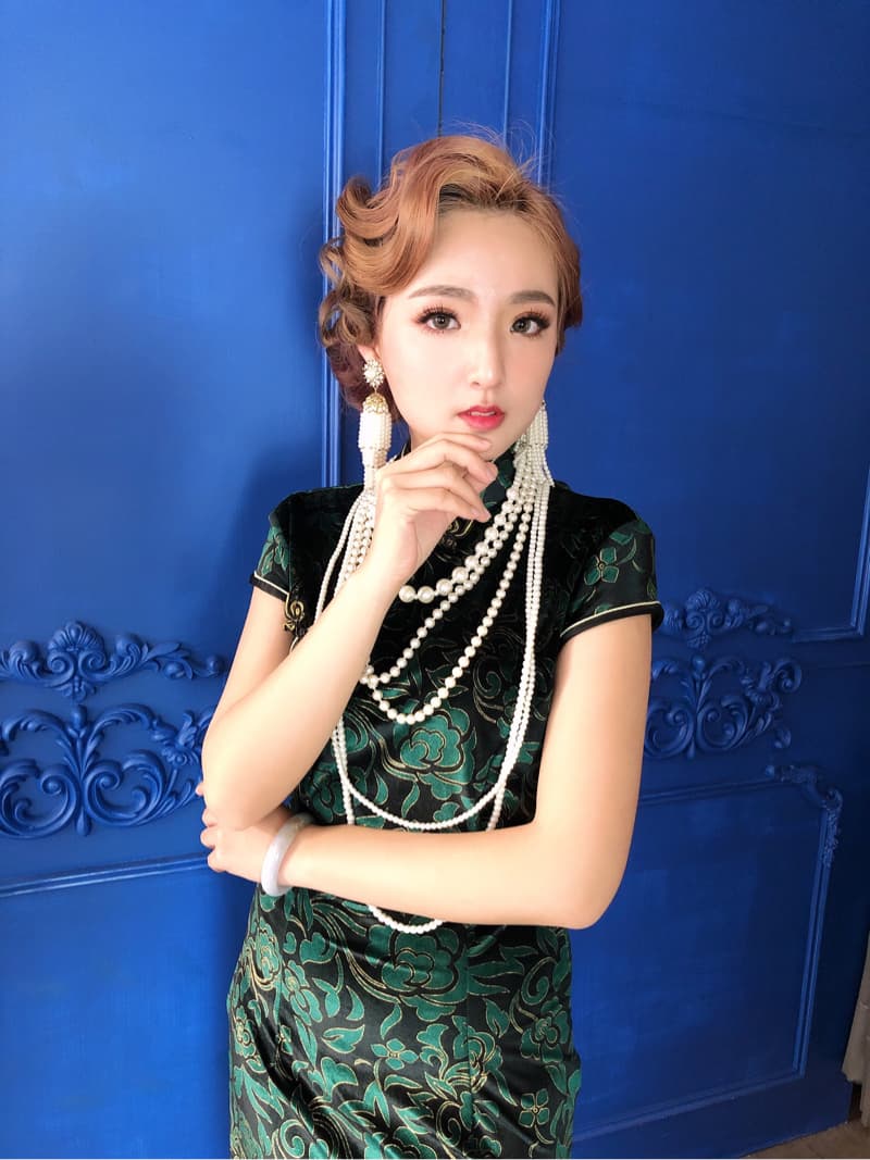這張「一位年輕女子身穿綠色旗袍，配戴珍珠項鍊和耳環，站在藍色背景前」的照片是。