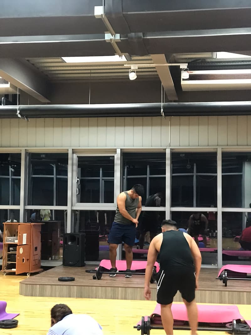 這張照片是一個健身房的場景，照片中最重要的是一個正在使用槓鈴的男人。他穿著黑色的背心和短褲，雙手握住槓鈴，準備開始運動。