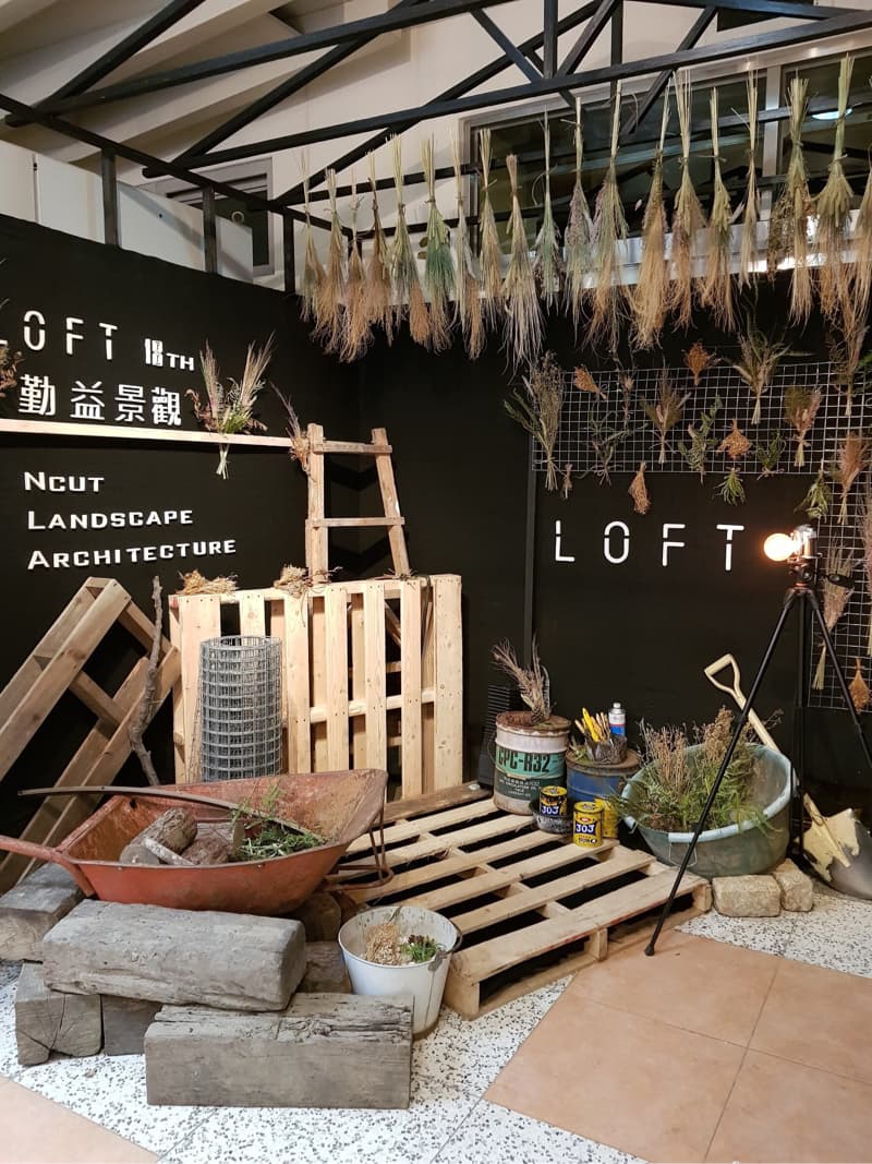 這張照片是一間木質裝潢的室內空間，空間裡擺放著許多木板、一個鐵製的輪子推車和一些盆栽，還有一隻梯子。牆上掛滿了乾草，呈現出自然樸實的風格。