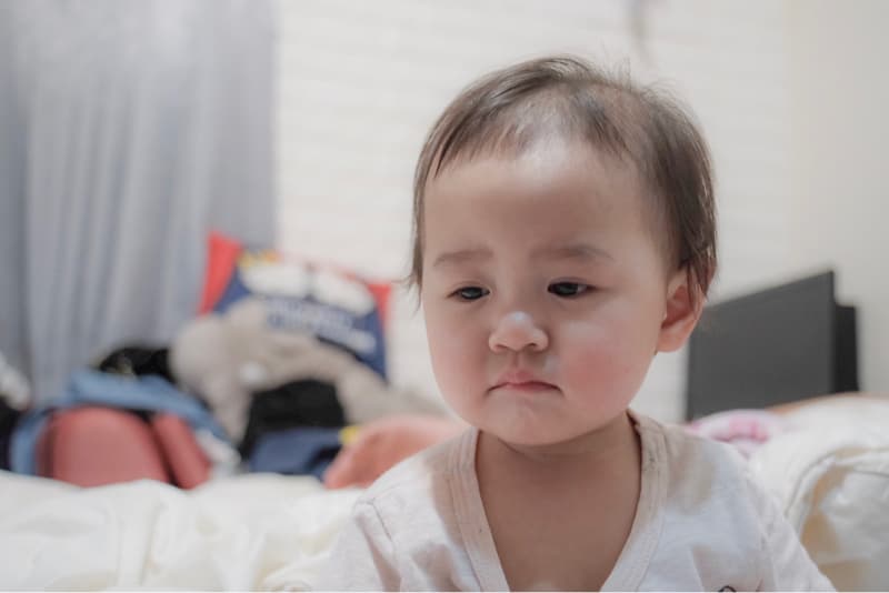 這張「嬰兒」的照片是一個穿著白色上衣的嬰兒，坐在床上，看著鏡頭，神情有點嚴肅。