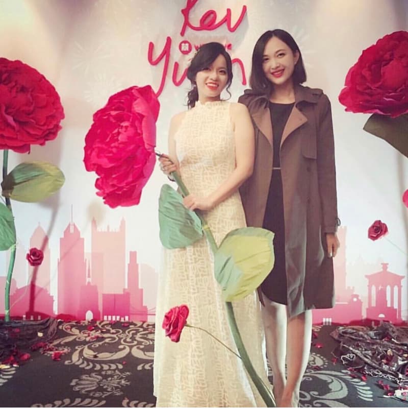 這張照片是兩個女生站在巨型玫瑰花前，她們都穿著漂亮衣服，背景是城市剪影和紅色的玫瑰花。