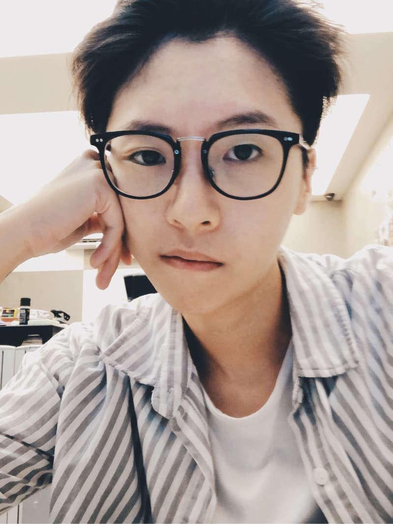 這張照片是一個年輕女子，她穿著一件白色底的灰色條紋襯衫，戴著黑色眼鏡，右手托著下巴，看起來有點憂鬱。