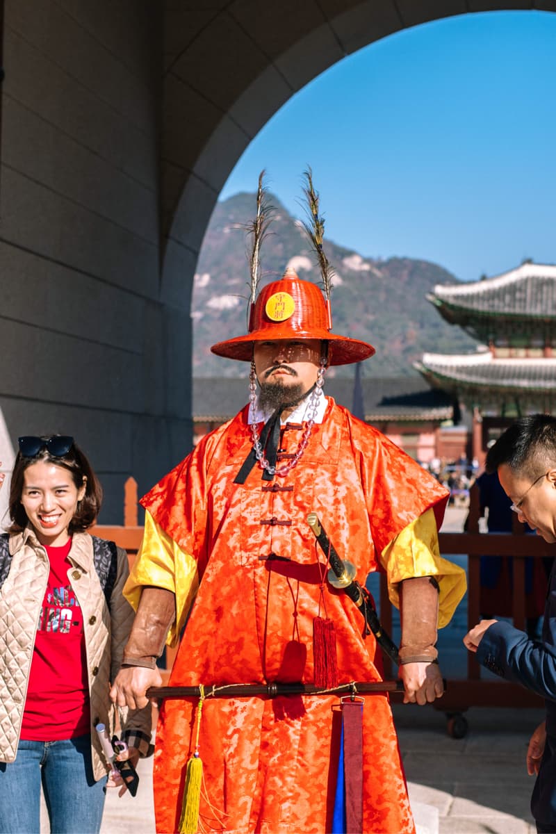這張「韓國傳統服飾」的照片是一個穿著鮮豔紅色長袍和紅色帽子，手持長劍的傳統韓國武士。他站在一個傳統的建築物前，表情嚴肅，看起來很有威嚴。