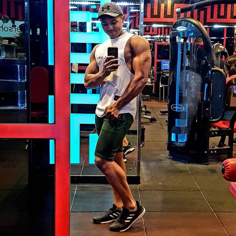 這張「健身房」的照片是一個男子在健身房自拍，他穿著白色背心和綠色運動短褲，戴著帽子，露出健壯的身材。