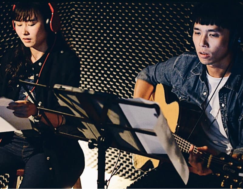 這張照片是兩個人在錄音室裡錄歌，他們正在認真地演唱，其中一位戴著耳機，另一位正在彈吉他，他們看起來很專注。