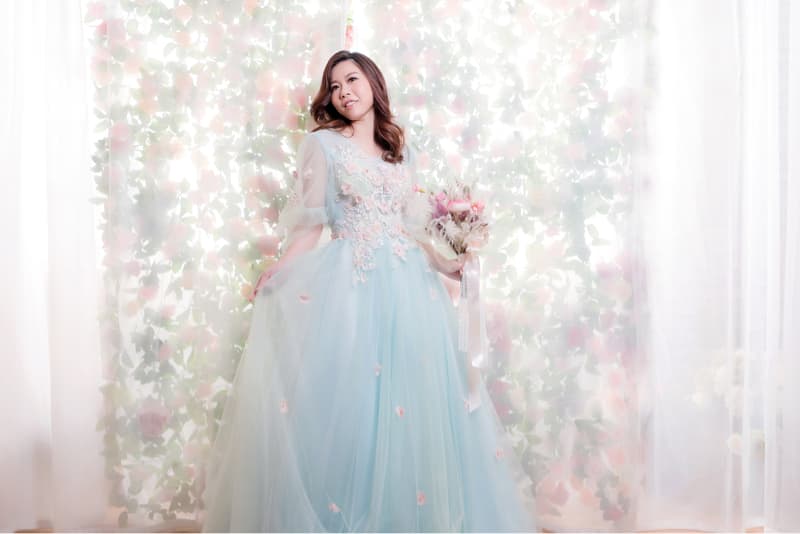 這張照片是一個穿著淡藍色禮服的女性，她手持花束，站在佈滿粉色花的背景前，並略帶微笑地看著鏡頭。