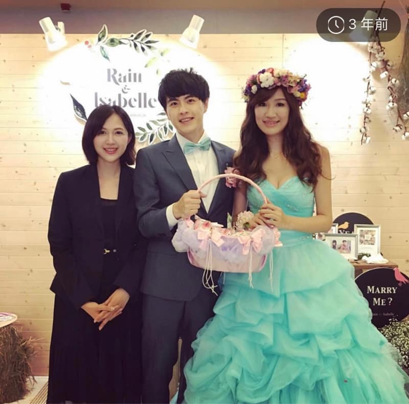 這張「婚禮」的照片是一對新人和一位女性友人，新人手拿著一個粉紅色的花籃，新娘穿著藍色的婚紗，新郎則穿著灰色的西裝。