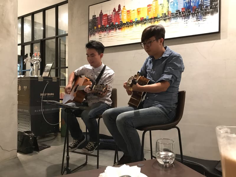 這張「兩個男生在餐廳彈吉他」的照片是，一個男生坐在椅子上，彈著木吉他，另一個男生坐在高腳椅上，彈著電吉他。