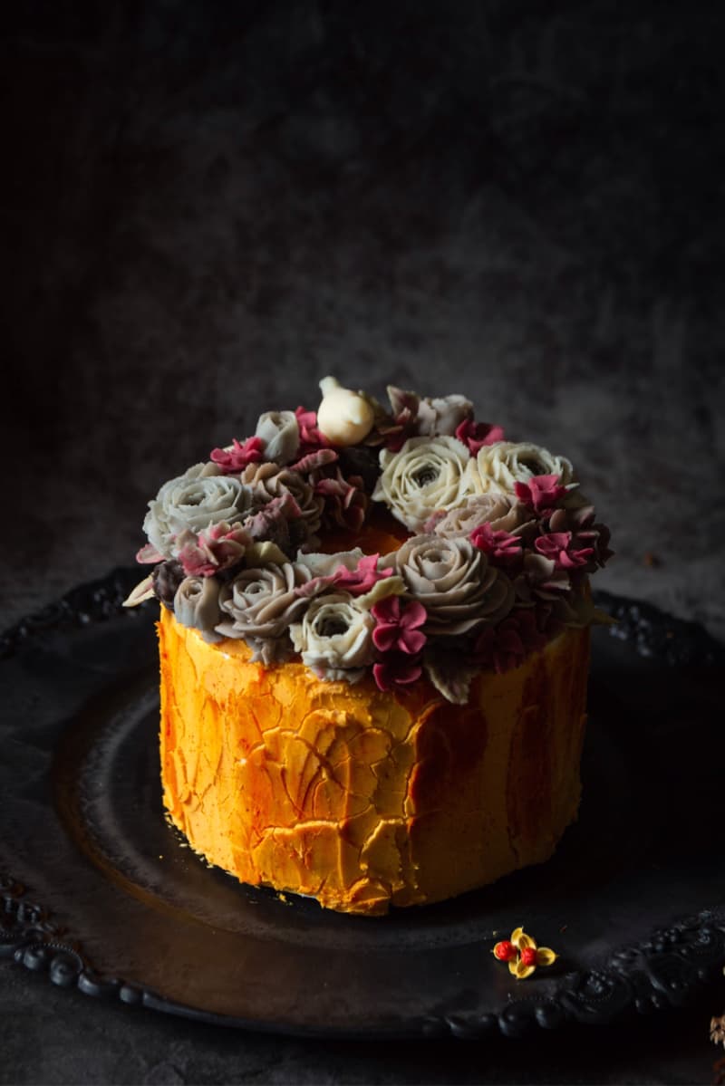 這張「蛋糕」的照片是一個以花卉裝飾的蛋糕，蛋糕是黃色的，上面放滿了白、粉紅色的花朵，背景是灰色的。