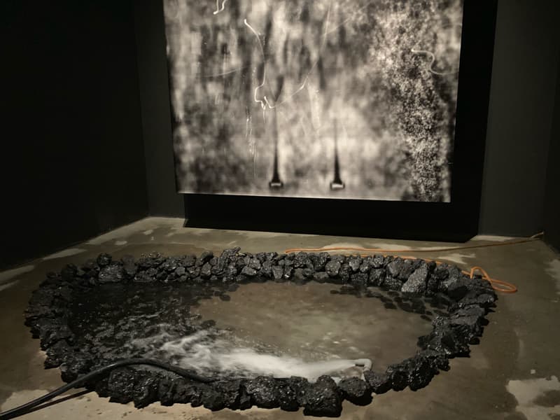 1) 照片展示了一個黑暗房間，房間中央有一條黑色的水管，水管在一個用黑石圍起來的圓形水池中央噴出水