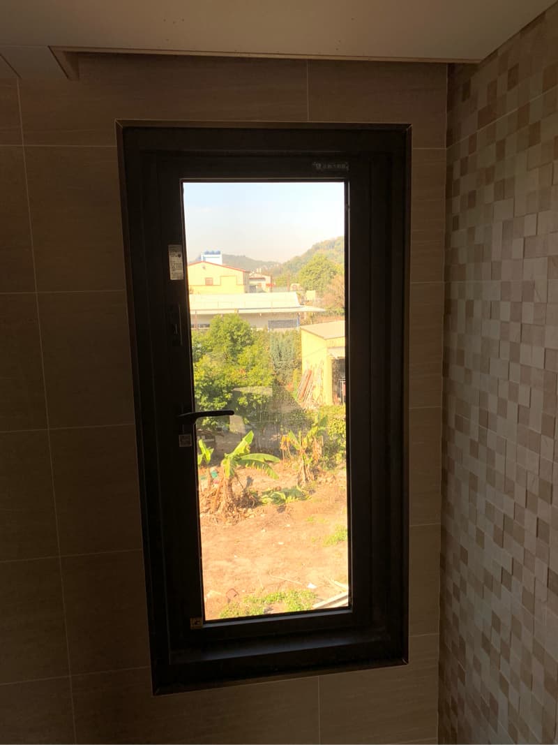 1) 照片顯示一扇黑色的窗戶，窗戶上有一隻黑色的門把， 窗戶外面可以看到一些樹木，一些建築物和遠處的綠色山丘