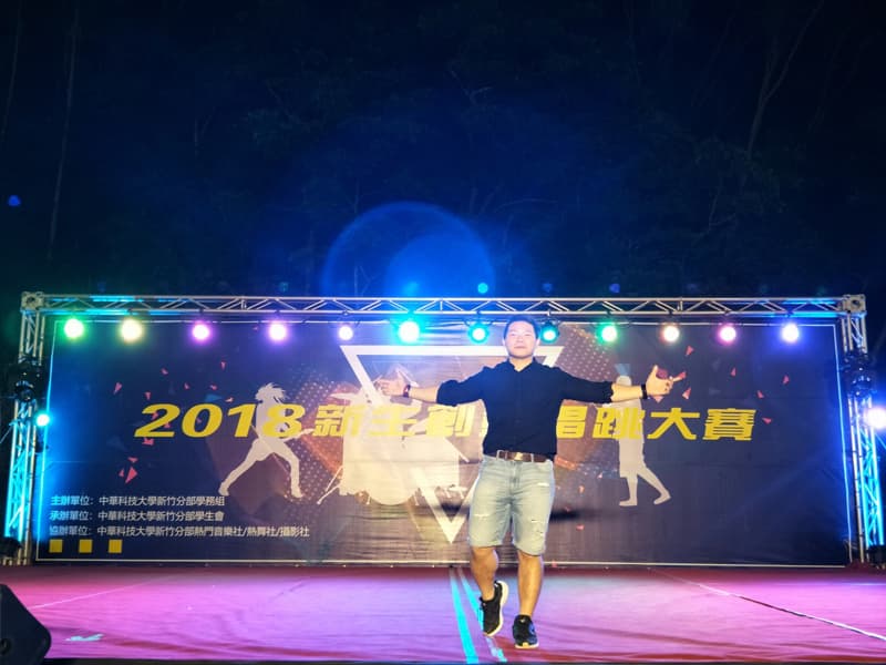 這張「舞臺表演」的照片是，一位男性站在舞臺上，背景是舞臺燈光與 2018 新生訓練的看板。