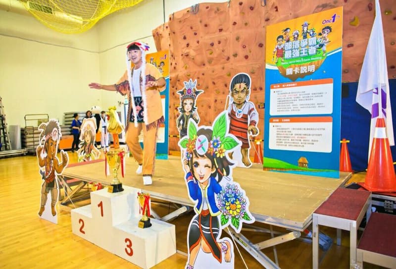 這張「原住民文化活動」的照片是，一個穿著傳統服裝的人在臺上表演，旁邊有獎盃和卡通人物。