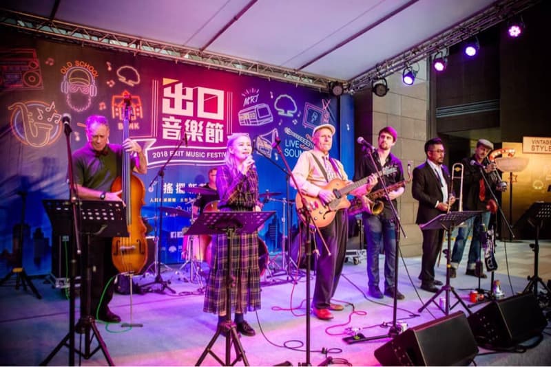 這張照片是一場戶外音樂表演，最重要的是舞台上的樂團，他們正在演奏音樂，樂團成員有主唱、吉他手、貝斯手、薩克斯風手和長號手。