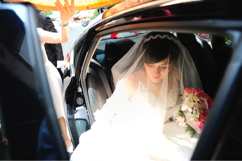 這張照片是一位穿著婚紗的新娘,她正準備坐進一輛黑色的汽車裡。她穿著白色的婚紗,頭戴頭紗,手裡拿著一束鮮花。