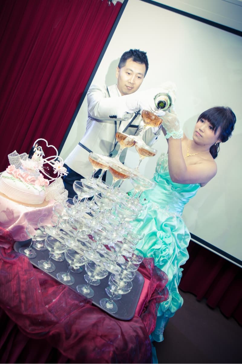 這張照片是一對新人在婚禮上舉行香檳塔儀式。新郎正在為香檳塔倒酒，新娘穿著藍色的禮服，站在一旁。他們面前擺放著一個精緻的蛋糕，看起來很浪漫。