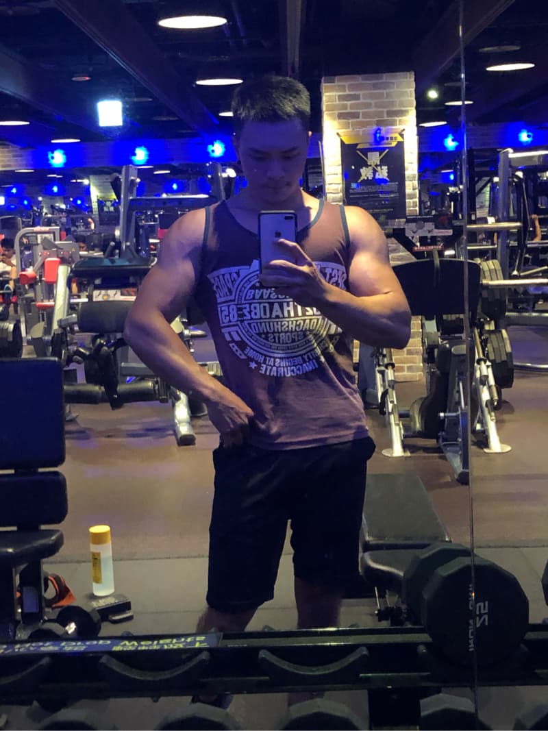 這張照片是一個在健身房裡自拍的年輕人，他穿著運動背心和短褲，顯得非常健壯，照片中他正對著鏡子，用手機自拍，背景是健身房的器材。