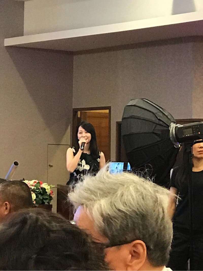 這張照片是一個女人在臺上講話，她拿著麥克風，穿著黑色的衣服，顯得很自信。