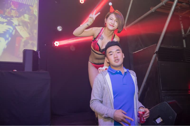 這張照片是一名男子和一名身穿紅色內衣的女子站在一起。男子戴著藍色帽子，穿著灰色外套和藍色襯衫，女子戴著紅色帽子，手放在男子肩膀上，做著勝利手勢。