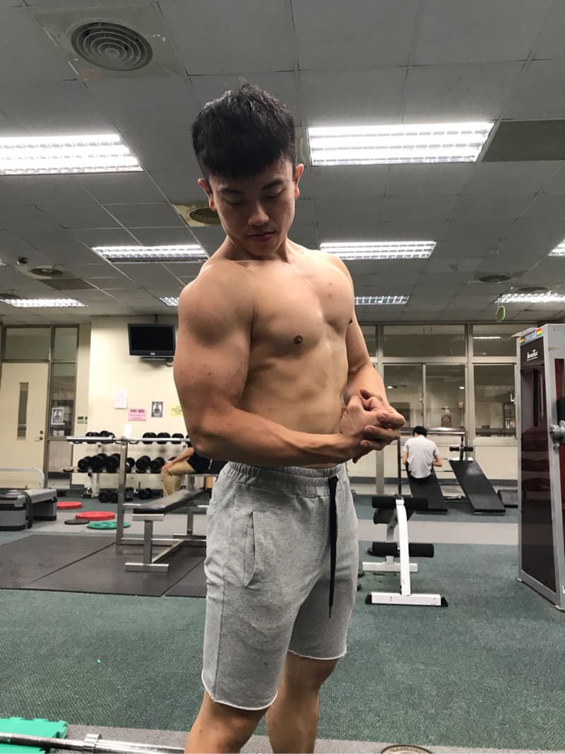 這張「健身房」的照片是一個年輕男子在健身房裡展示他強壯的肌肉，他穿著灰色運動短褲，背景是健身房的器材。