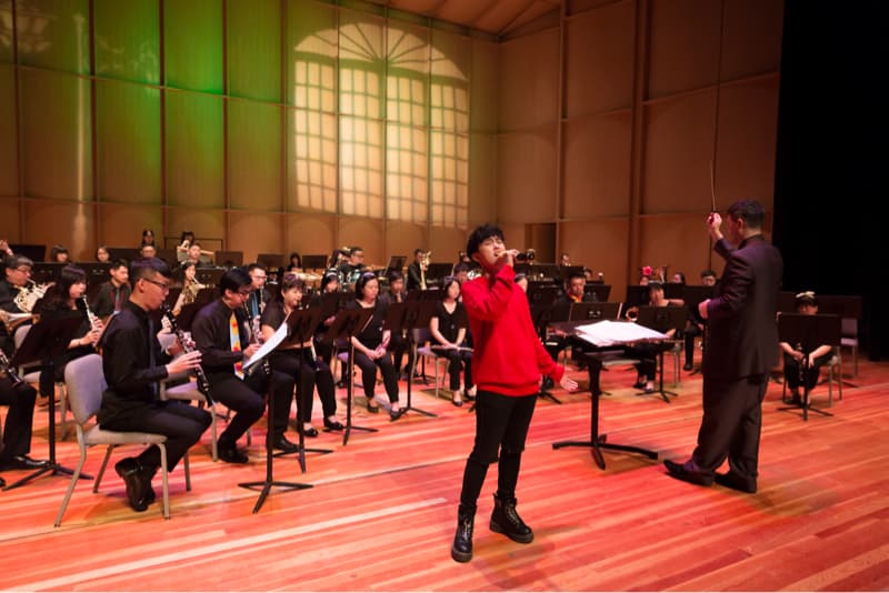 這張照片是正在演唱的歌手，他身穿紅色上衣，站在舞台中央，指揮則在後方揮舞著指揮棒。