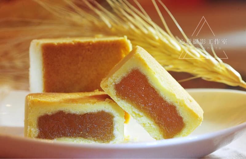 這張「鳳梨酥」的照片是三塊鳳梨酥放在白色盤子上，其中一塊鳳梨酥被切開，露出裡面的鳳梨餡。旁邊有一根麥穗，讓照片更有質感。