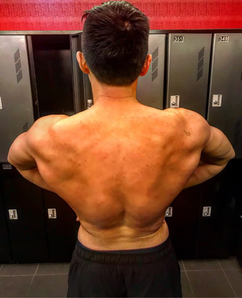 這張「一個男人在更衣室裡」的照片是，他赤裸上身，展示出他強壯的背部肌肉。他站在一個排滿黑色儲物櫃的牆壁前。