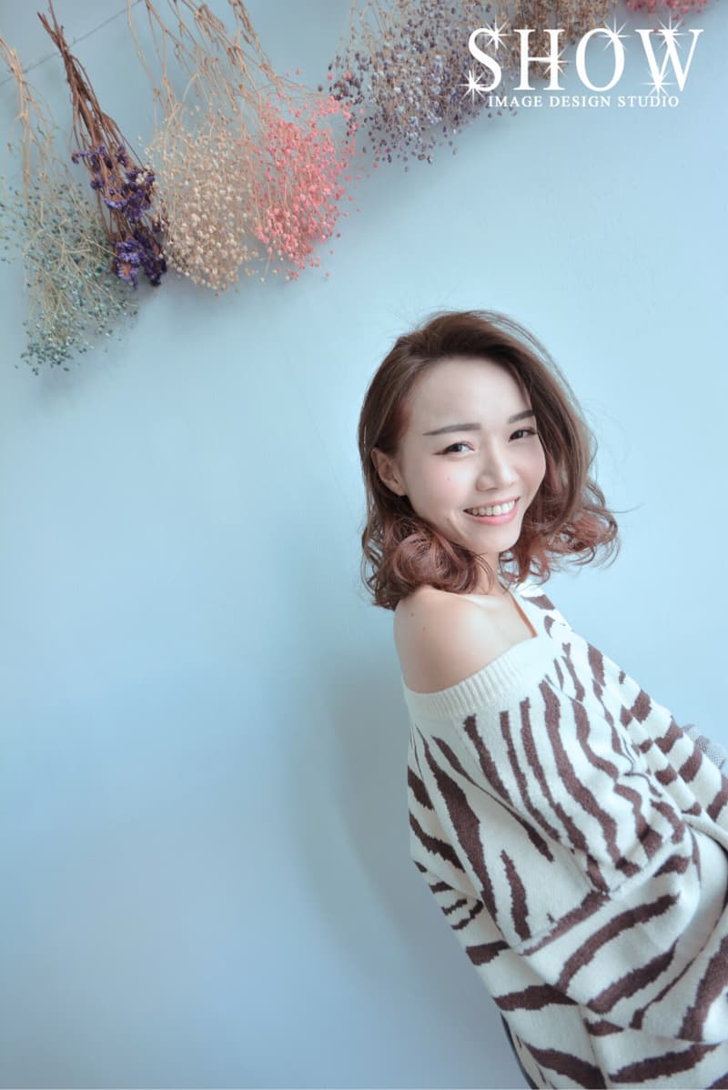 這張「SHOW IMAGE DESIGN STUDIO」的照片是一個年輕女子，她穿著一件棕色和白色條紋毛衣，笑容燦爛，背景是淺藍色牆壁和一些乾燥花朵。