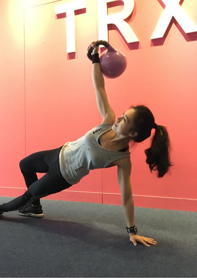 這張照片是一個年輕女性在健身房裡做運動。她正在舉起一個粉紅色的壺鈴，她的動作看起來很吃力，但她的眼神很堅定，顯示出她對自己的目標充滿信心。