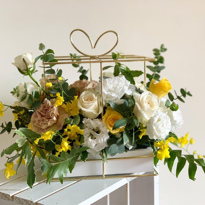 這張照片是一個用金屬框架做成的禮盒，裡面裝滿了各種顏色鮮豔的花朵，包括白色、黃色和粉色。花朵佈滿整個盒子，看起來十分繽紛漂亮。