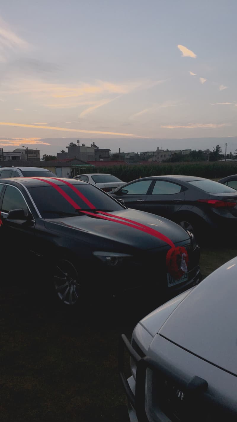 這張照片是一輛黑色轎車，車頭綁著兩條紅色緞帶，並繫上一個紅色花球，顯得喜氣洋洋。